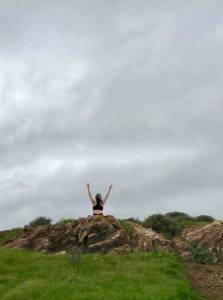 Photo of woman sitting atop a grassy hill, arms skyward