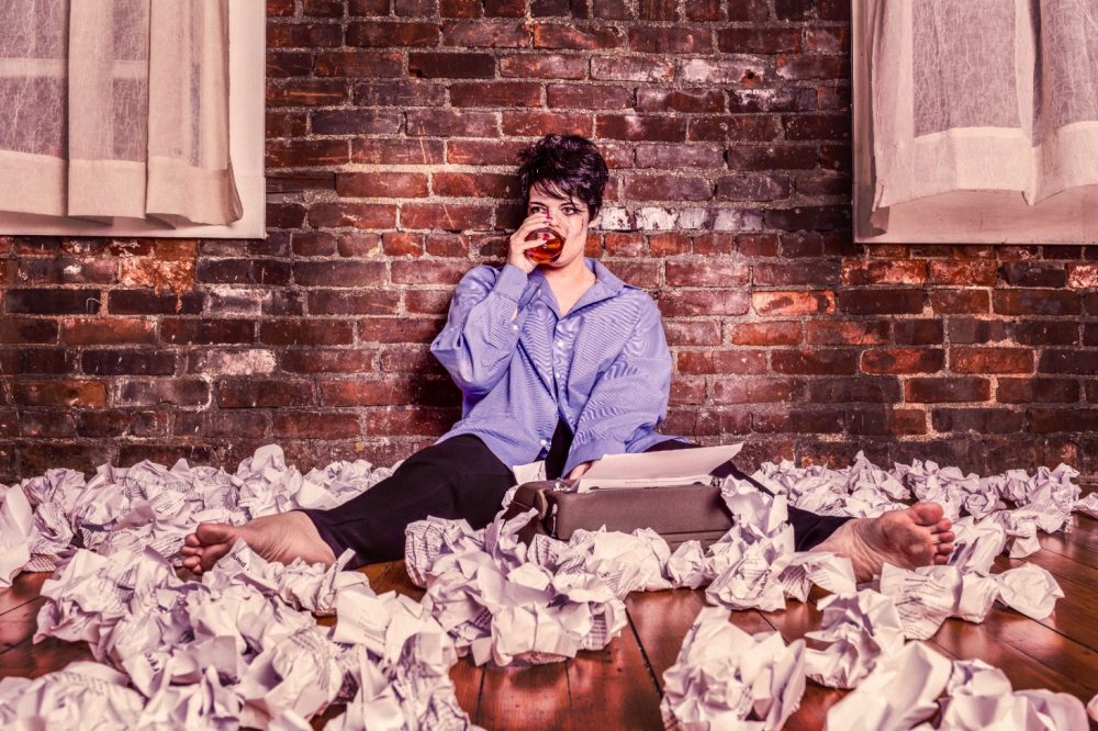 Photo of woman writer sitting in front of a typewriter, amid many crumpled pages, drinking alcohol