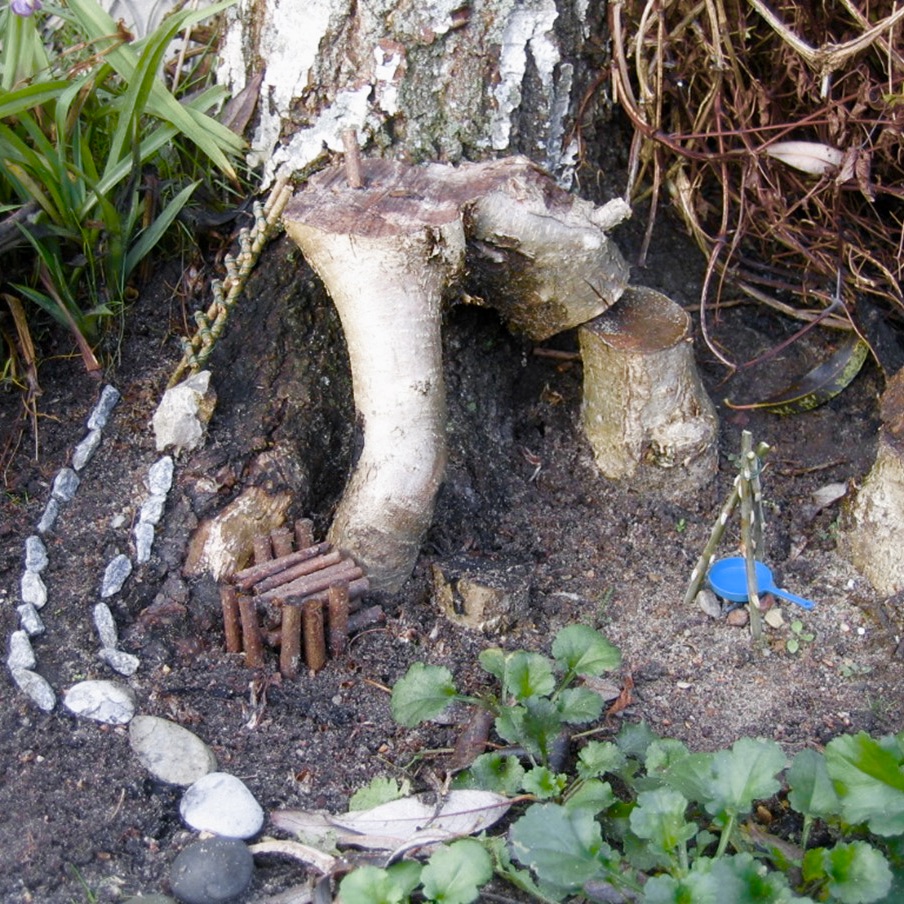 Photo of Birgit's elfish abode under a tree in her garden, replete with small rocks to mark a path, and doll-sized pots, pans, and chairs.