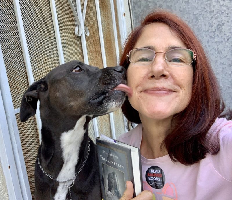 Photo of K-D, a black and white pitbull mix dog, gives da-AL a kiss on the cheek as she holds a book.