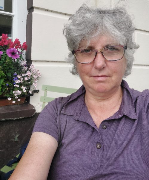 Selfie by Chichester, UK, taken late August 2024 in Lampeter, on a cycling holiday in Wales. She's dressed in a purple shirt and is sitting near a pot of flowers.
