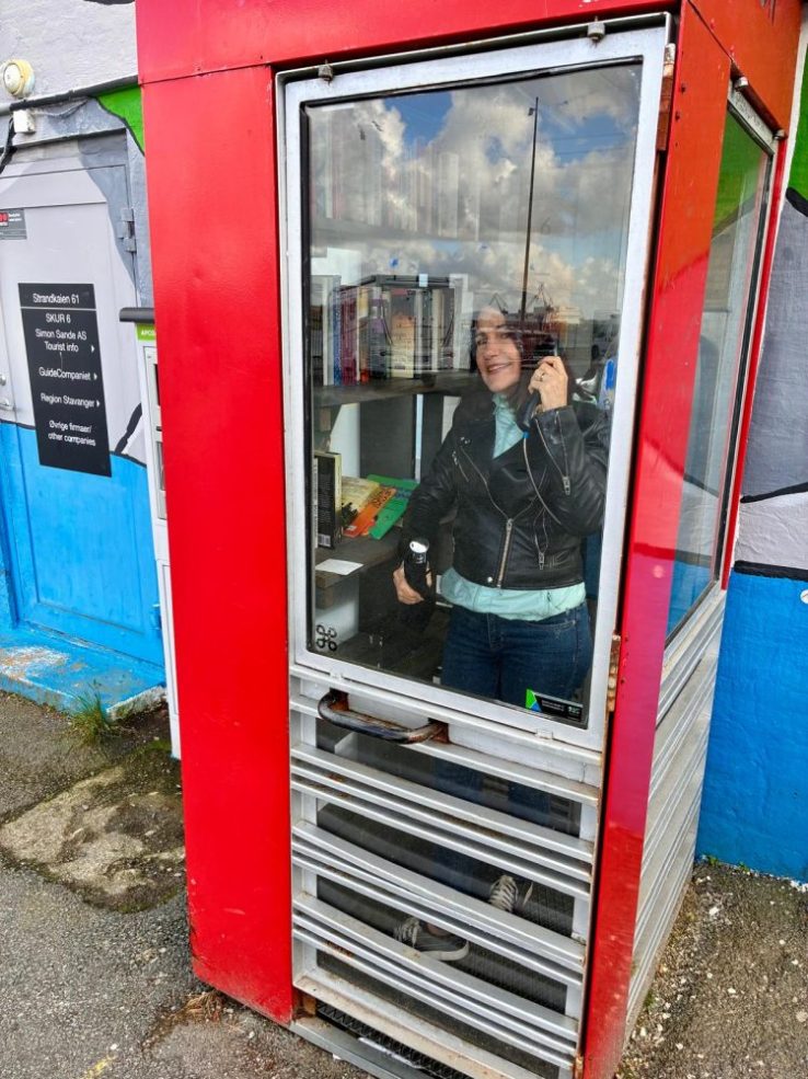 da-AL pretends to make a phone call from an old red telephone booth that's lined with books on shelves.