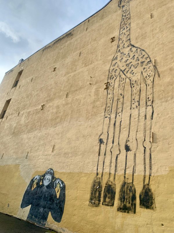 Mural on the side of the building of a monkey plugging its ears with its fingers, a giraffe towering over it.